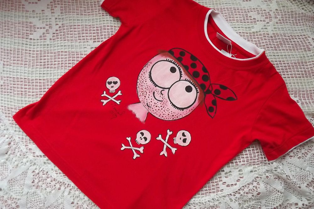 Veselý pirát namalovaný na červeném tričku s krátkým rukávem a vrstveným efektem, kolem piráta jsou lebčičky se zkříženými hnáty. veliksot 128